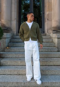 Cárdigan con cremallera verde oliva de textura acanalada, camiseta blanca, pantalones blancos de pierna ancha y zapatillas blancas con detalles negros, de pie sobre escalones de piedra.