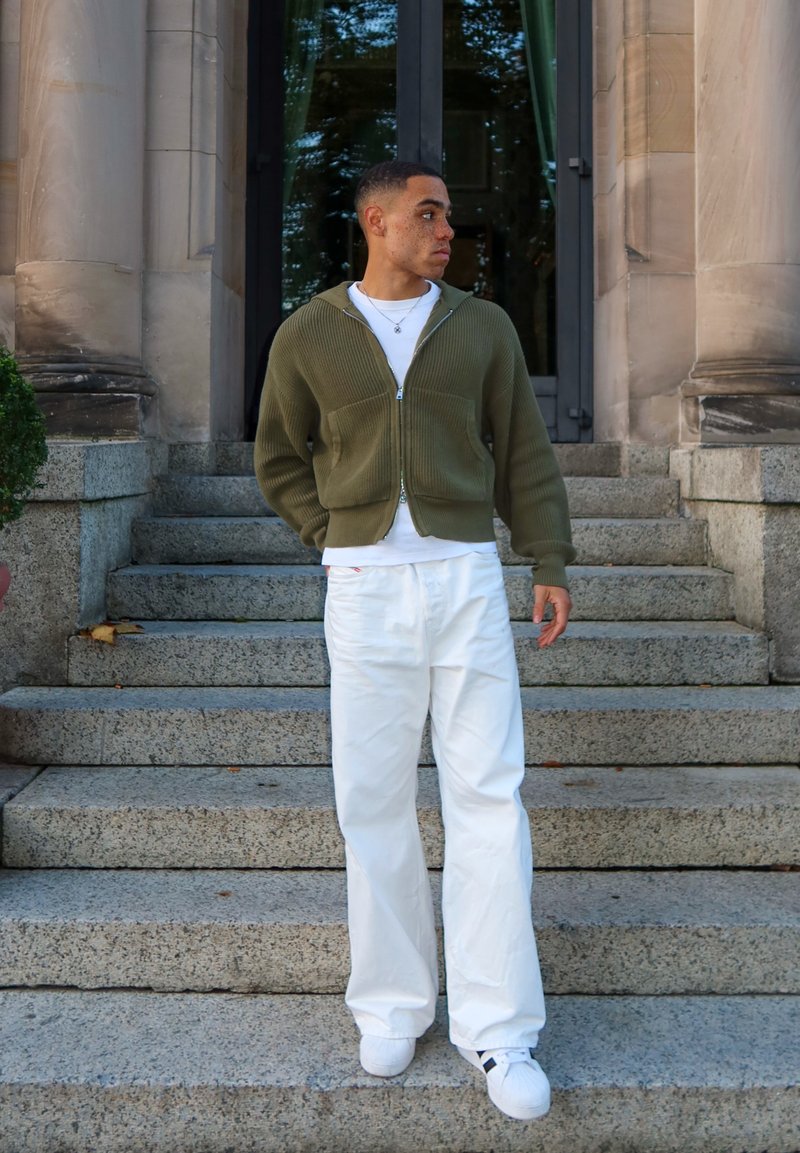 Olivovozelený kardigán so zipsom a žíhanou textúrou, biele tričko, biele široké nohavice a biele tenisky s čiernymi akcentmi, stojaca na kamenných schodoch.