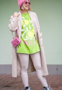 Πράσινο γραφικό t-shirt με σχέδιο λεμονιού, μπεζ παλτό, εφαρμοστά μπεζ κολάν, ροζ σκουφί και τσάντα. Η υπόδηση περιλαμβάνει μαύρα αθλητικά παπούτσια.