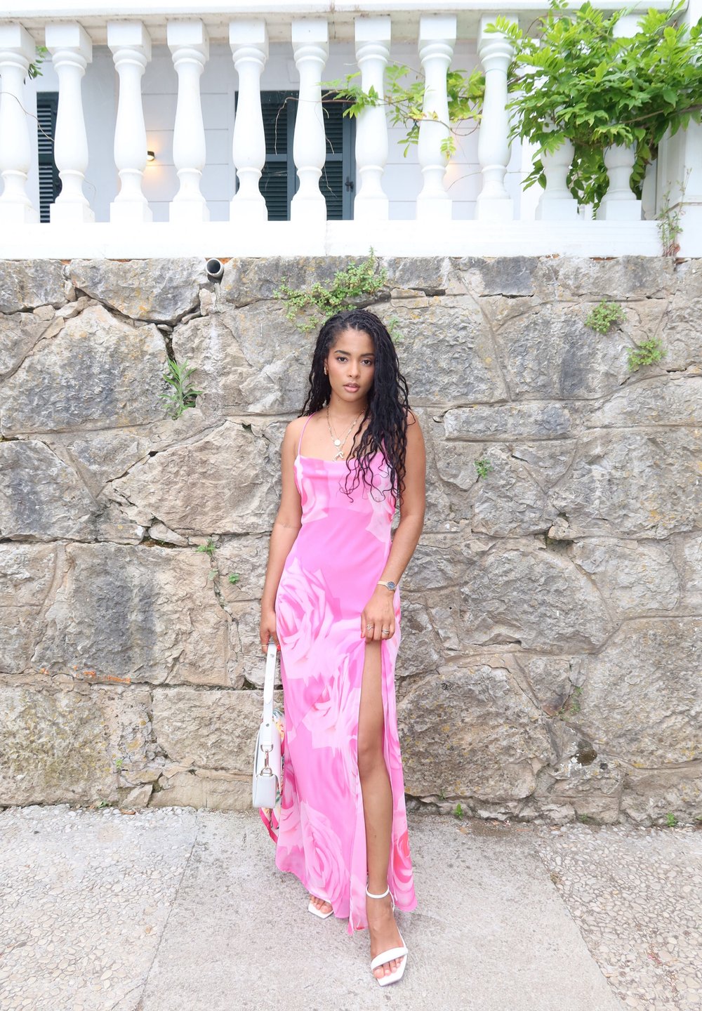 Rosa blommig maxikjol med en hög slits och tunna axelband. Ett par vita klacksandaler och en liten vit handväska. Texturerad stenvägg i bakgrunden.