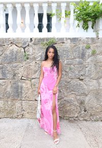 Rosa blommig maxikjol med en hög slits och tunna axelband. Ett par vita klacksandaler och en liten vit handväska. Texturerad stenvägg i bakgrunden.