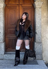 Giovane donna che indossa una giacca di pelle con colletto in pelliccia, stivali neri, gambaletti bianchi, una gonna di jeans e tiene una borsa nera, pose accanto a una porta di legno.