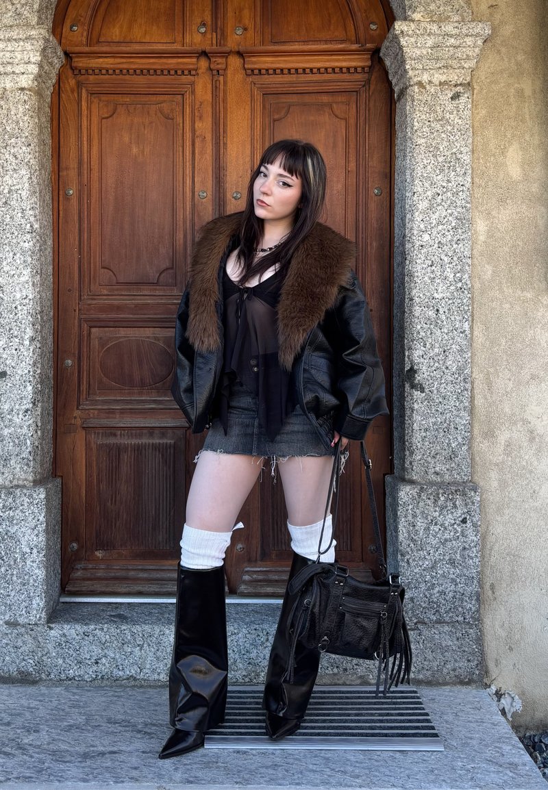 Junge Frau in einer Lederjacke mit Pelzkragen, schwarzen Stiefeln, weißen Schenkelwärmern, einem Denim-Rock und einer schwarzen Handtasche, posiert vor einer Holztür.