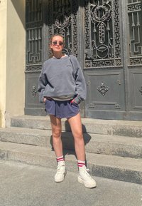 Sweatshirt cinza oversized, calções azul-marinho, ténis brancos de cano alto e meias risca vermelha. De pé em degraus de pedra com portas de metal decorativas.