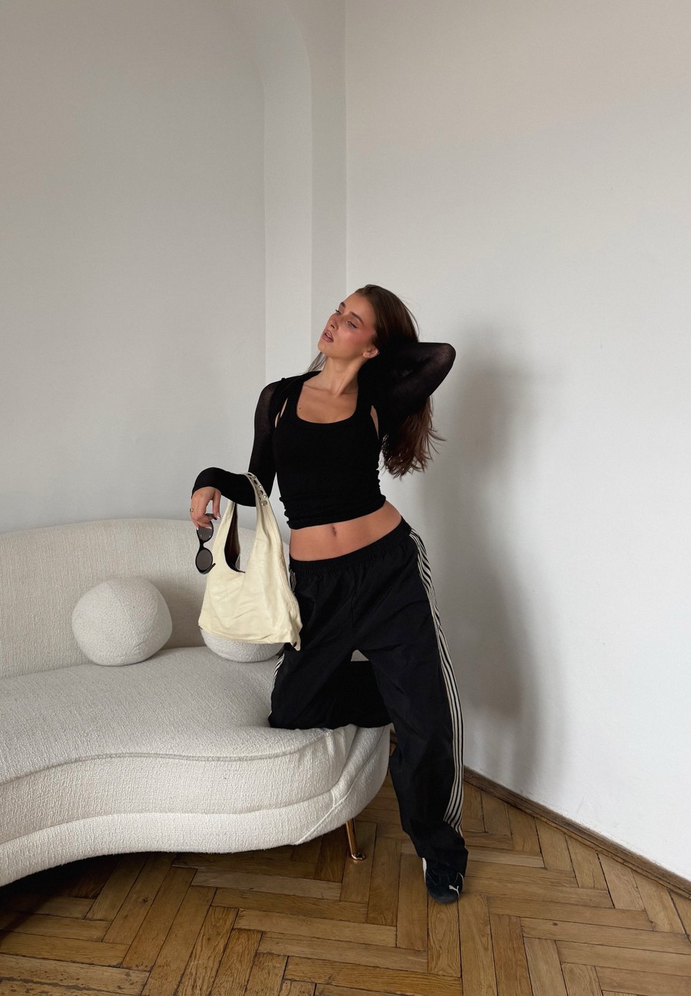 Czarny krótki top i luźne czarne spodnie dresowe z białymi bocznymi paskami; trzyma kremową torebkę na ramię i czarne okulary przeciwsłoneczne.