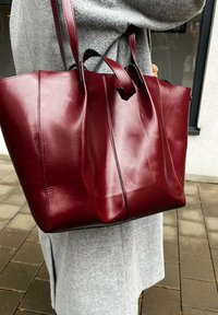 Borsa tote in pelle bordeaux con forma strutturata, due manici, cuciture pulite e finitura lucida; appoggiata su un cappotto grigio.