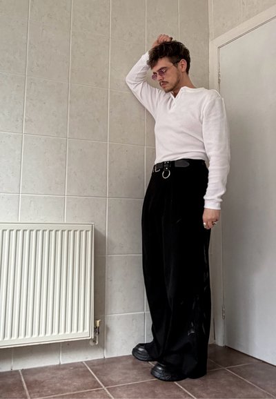 Persona con camisa blanca de manga larga, pantalones negros de talle alto y gafas de sol de pie en una habitación con azulejos, apoyando la mano en la cabeza.