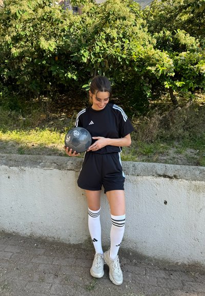 Conjunto deportivo negro con rayas blancas, camiseta ajustada y pantalones cortos, calcetas blancas hasta la rodilla con tres rayas negras, y balón deportivo gris.