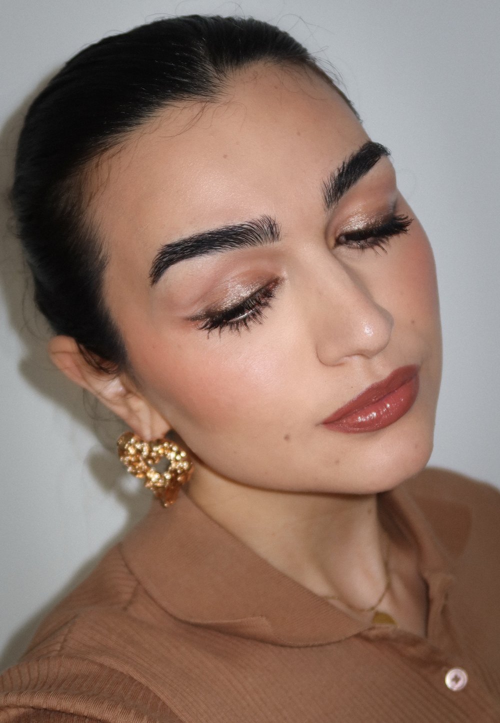 Eine Nahaufnahme einer Person mit dunklem Haar, die große, goldene Herzohrringe trägt. Das Outfit besteht aus einem hellbraunen Hemd mit Kragen und texturiertem Finish.