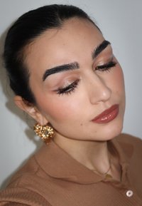 Eine Nahaufnahme einer Person mit dunklem Haar, die große, goldene Herzohrringe trägt. Das Outfit besteht aus einem hellbraunen Hemd mit Kragen und texturiertem Finish.
