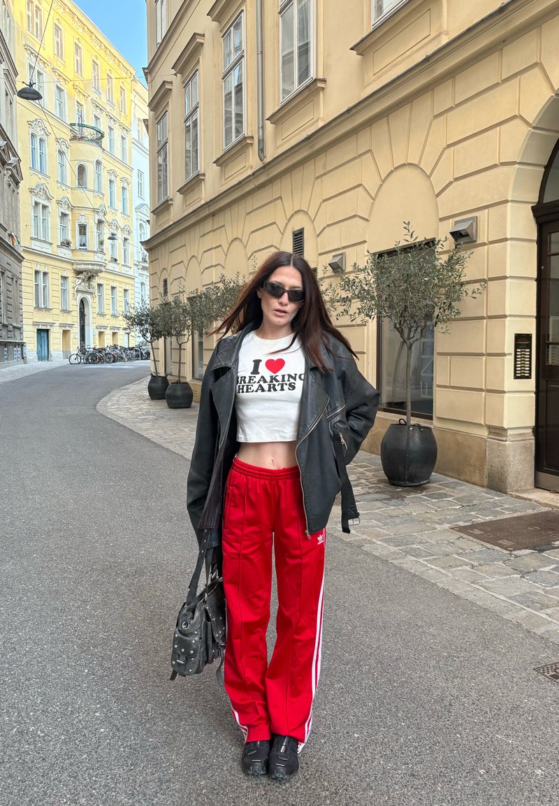 Žena hoda gradskom ulicom noseći sunčane naočale, crnu kožnu jaknu, bijeli kratki top s natpisom, crvene trenirke i crne cipele, držeći torbu.