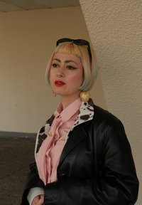 Vrouw met kort pastelkleurig haar, rode lippenstift, hartvormige gouden oorbellen, roze blouse met een koeienprint kraag, zwarte leren jack, en een zonnebril op het hoofd.