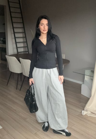 Sudadera negra con cremallera y mangas largas, pantalones de chándal grises de pierna ancha, bolso negro y zapatillas negras. Entorno interior con suelo de madera y decoración clara.