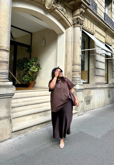 Camiseta oversized marrón y una falda larga marrón, combinadas con zapatos claros. De pie delante de un edificio con columnas y una planta en maceta.