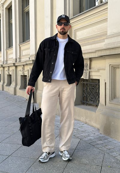 Hombre con chaqueta negra, camisa blanca, pantalones beige, zapatillas deportivas, gafas de sol y gorra, sosteniendo una bolsa negra, de pie en la acera de la ciudad.