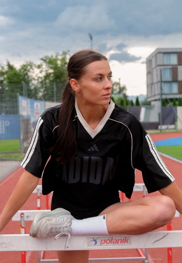 Athlète féminine en tenue de sport noire étire sa jambe sur une haie sur une piste extérieure pendant une journée nuageuse, regardant sur le côté.