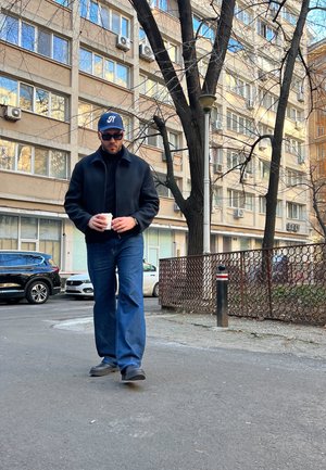 Chaqueta negra, suéter de cuello alto negro, jeans azules y gafas de sol, usado por una persona que sostiene una taza de café mientras camina por una calle de la ciudad.