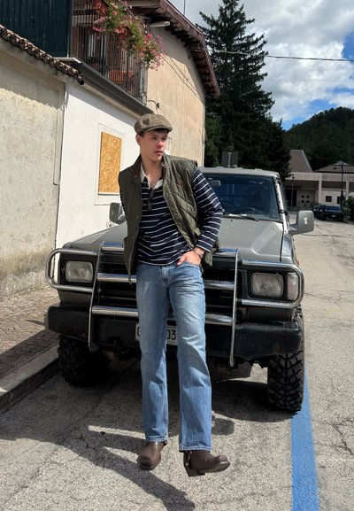 Chaleco acolchado verde sobre una camisa de rayas azul marino y blanca de manga larga, vaqueros azul claro y botas de cuero marrón. SUV aparcado de fondo.