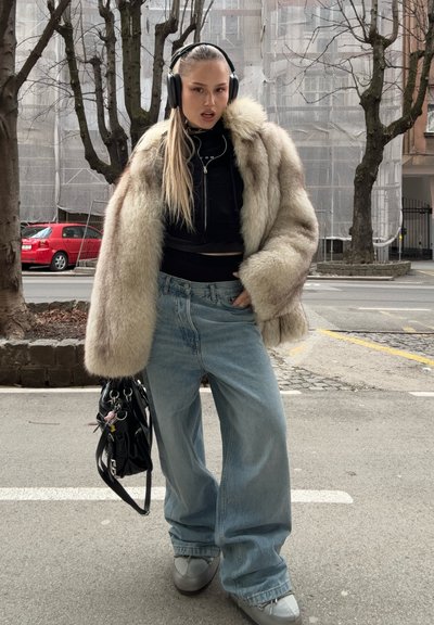 Abrigo de piel sintética con tonos marrón claro y crema, top corto negro, jeans sueltos azules, zapatos grises sin cordones y un bolso negro.