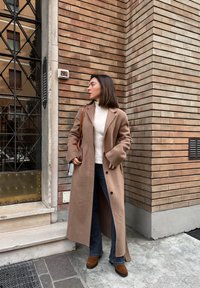 Long manteau en laine beige, col roulé crème, jean évasé foncé et bottes marron à talons. Debout contre un mur en brique près d'une porte.
