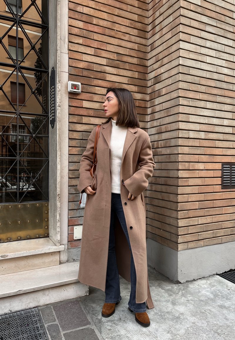 Long beige wool overcoat, cream roll-neck jumper, dark flared jeans, and brown heeled boots. Standing against a brick wall near a door.
