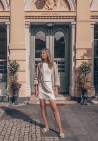 Μοντέλο που φοράει ένα ανοιχτό γκρι πουκάμισο από λινό πάνω από ένα λευκό φόρεμα, με ανοιχτού γκρι σανδάλια. Το φόντο περιλαμβάνει πράσινες διπλές πόρτες και γλάστρες με φυτά.