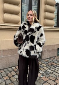 Une femme blonde se tient sur une rue pavée, portant un manteau en fausse fourrure noir et blanc à pois, un pantalon large noir, tenant un petit sac à main marron près d'un bâtiment beige.