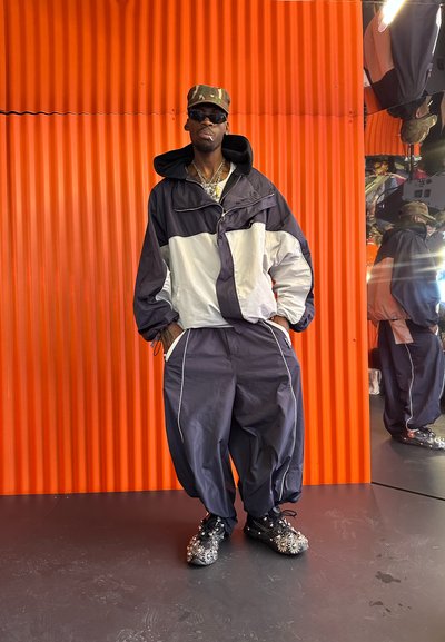 Chaqueta cortaviento en navy y gris con acentos reflectantes, combinada con pantalones holgados y zapatillas negras adornadas. Fondo naranja.