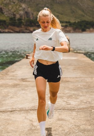T-shirt bianca Adidas con logo verde, pantaloni corti neri con strisce bianche, che corre su un molo di cemento vicino all'acqua, indossando un orologio e sneakers.