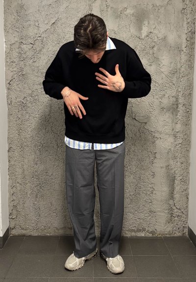 Sudadera negra sobre una camisa a rayas azul y blanca, pantalones grises y zapatillas de deporte gris claro. Fondo de pared de concreto.