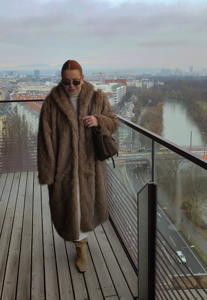 Manteau long en fausse fourrure marron, coupe oversize, porté sur une tenue de couleur claire. Complété par des lunettes de soleil rondes et un sac marron.