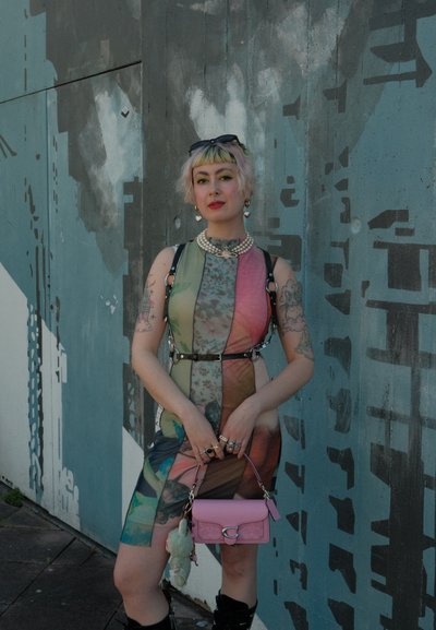 Vestido multicolorido con diseño de patchwork, corte sin mangas y silueta ajustada. Los accesorios incluyen un bolso rosa y múltiples anillos.