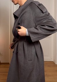 Trench-coat ceinturé en laine grise avec un large col, des détails à boutons sur les épaules, et une finition texturée, porté sur une chemise claire.