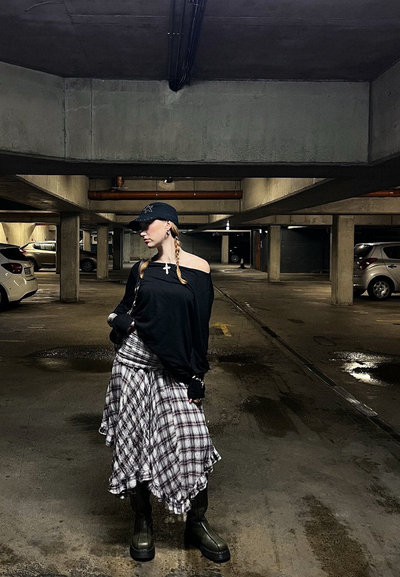 Femme aux cheveux tressés portant une casquette noire, un haut noir décolleté aux épaules, une jupe à carreaux et des bottes, se tient dans un parking en béton faiblement éclairé.