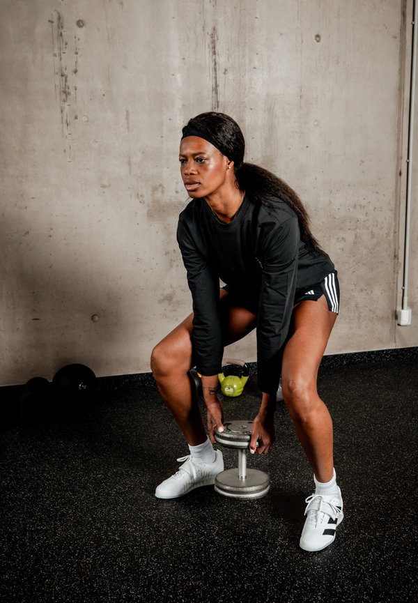 Kvinde i sort langærmet trøje og shorts løfter en håndvægt i et fitnesscenter med sort gummigulv og betonvægge.