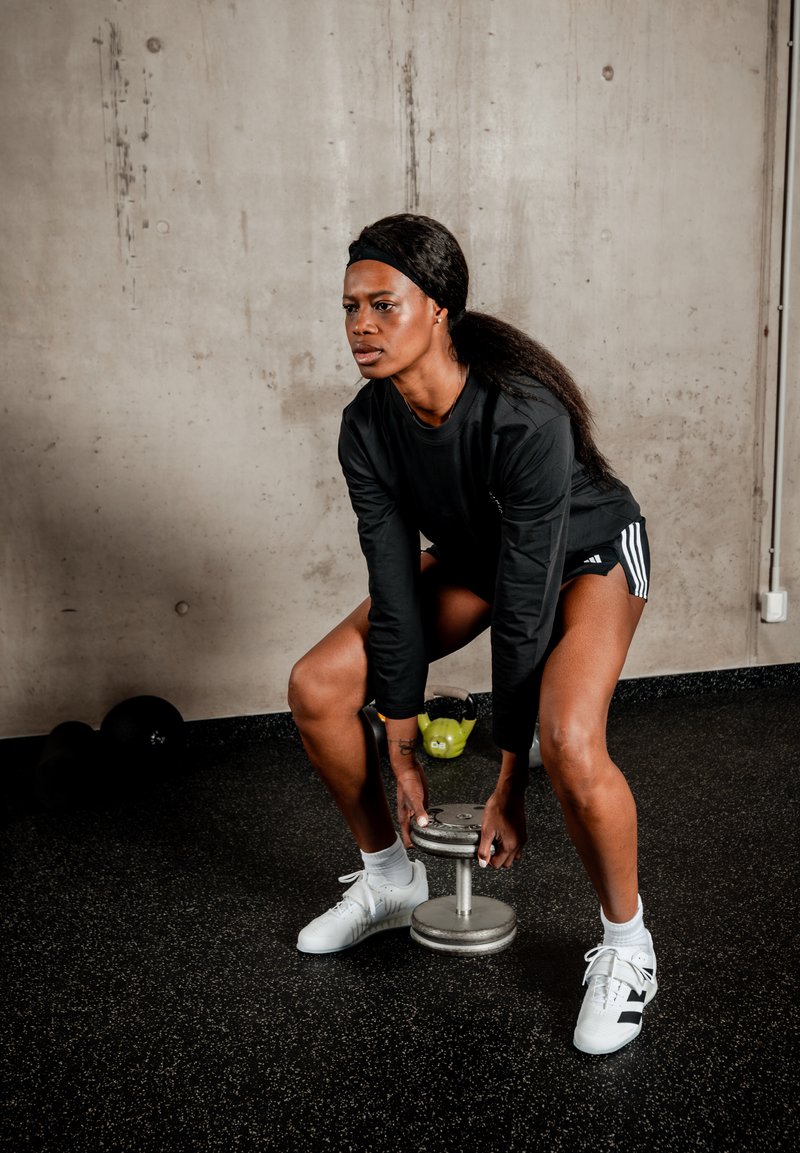 Femme en manches longues noires et short soulevant un haltère dans une salle de sport avec un sol en caoutchouc noir et des murs en béton.