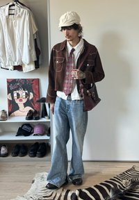 Homem a usar uma jaqueta castanha, camisa branca, gravata estampada, jeans largos azul claro, boné branco, e a carregar uma mala de ombro bordô numa divisão com roupas e sapatos.