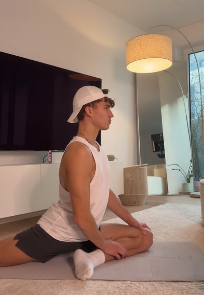 Joven con camiseta sin mangas blanca y gorra estira sobre una esterilla de yoga en una sala de estar con una lámpara de pie y un televisor grande.