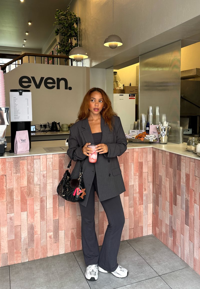 Grigio blazer oversize e top nero abbinati a pantaloni scuri, mentre tiene una bevanda rosa, davanti a un bancone in piastrelle rosa chiaro in un caffè.