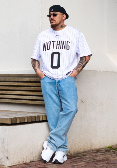 Hombre con brazos tatuados que lleva gafas de sol, boina negra, camiseta blanca "NOTHING 0", jeans azules y zapatillas blancas apoyado contra una pared cerca de un banco de madera.