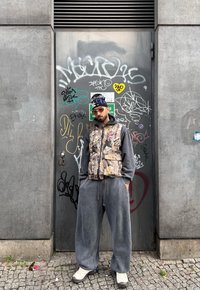 Homme portant un gilet camouflage sur un sweat à capuche gris, un pantalon gris ample, des baskets blanches, des lunettes de soleil orange et un bonnet noir, debout contre une porte en métal couverte de graffiti.