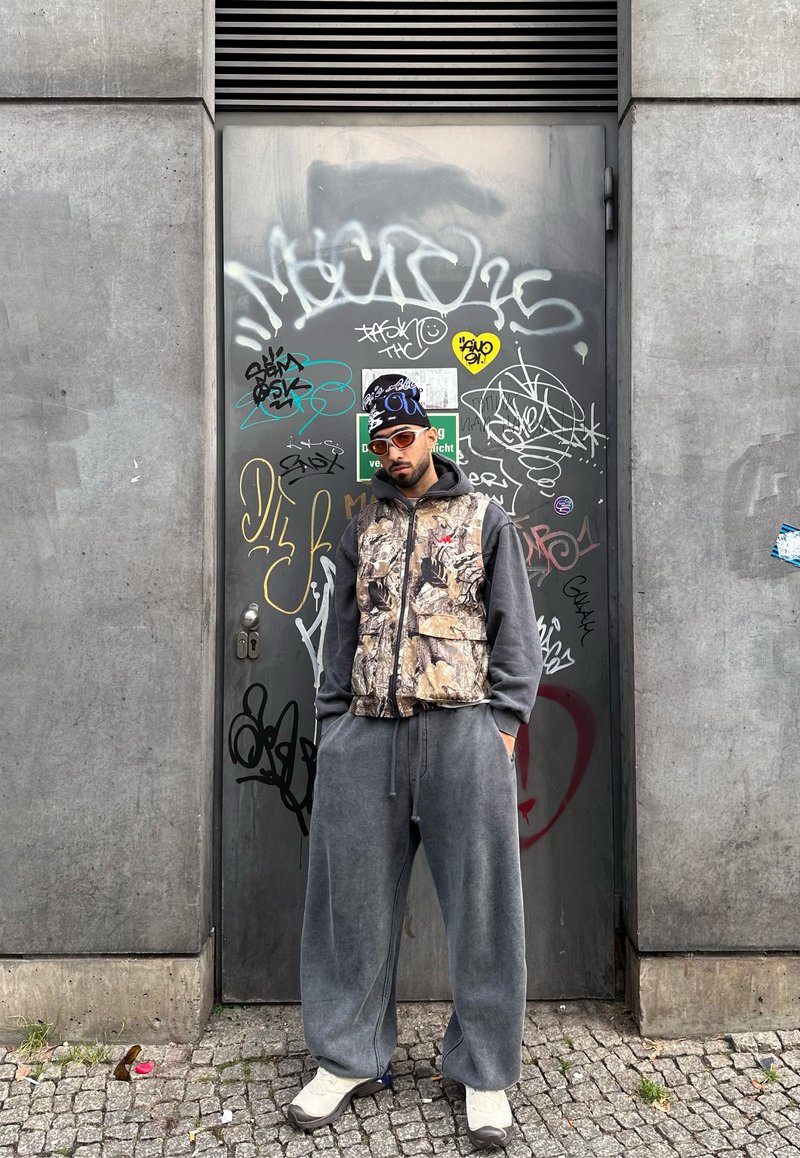 Homme portant un gilet camouflage sur un sweat à capuche gris, un pantalon gris ample, des baskets blanches, des lunettes de soleil orange et un bonnet noir, debout contre une porte en métal couverte de graffiti.