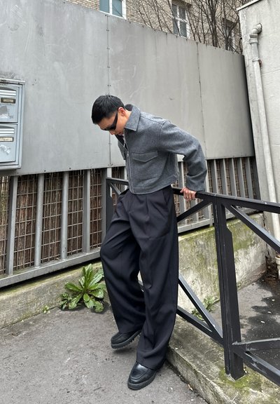 Hombre con chaqueta gris y pantalones anchos negros apoyado en una barandilla de metal negra en una acera urbana, con una pared gris y buzones detrás.