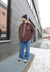 Strukturert, rustfarvet jakke med forlommer, lag på en brun og hvidstribet skjorte. Løse blå jeans med hvide detaljer. Sorte sneakers.