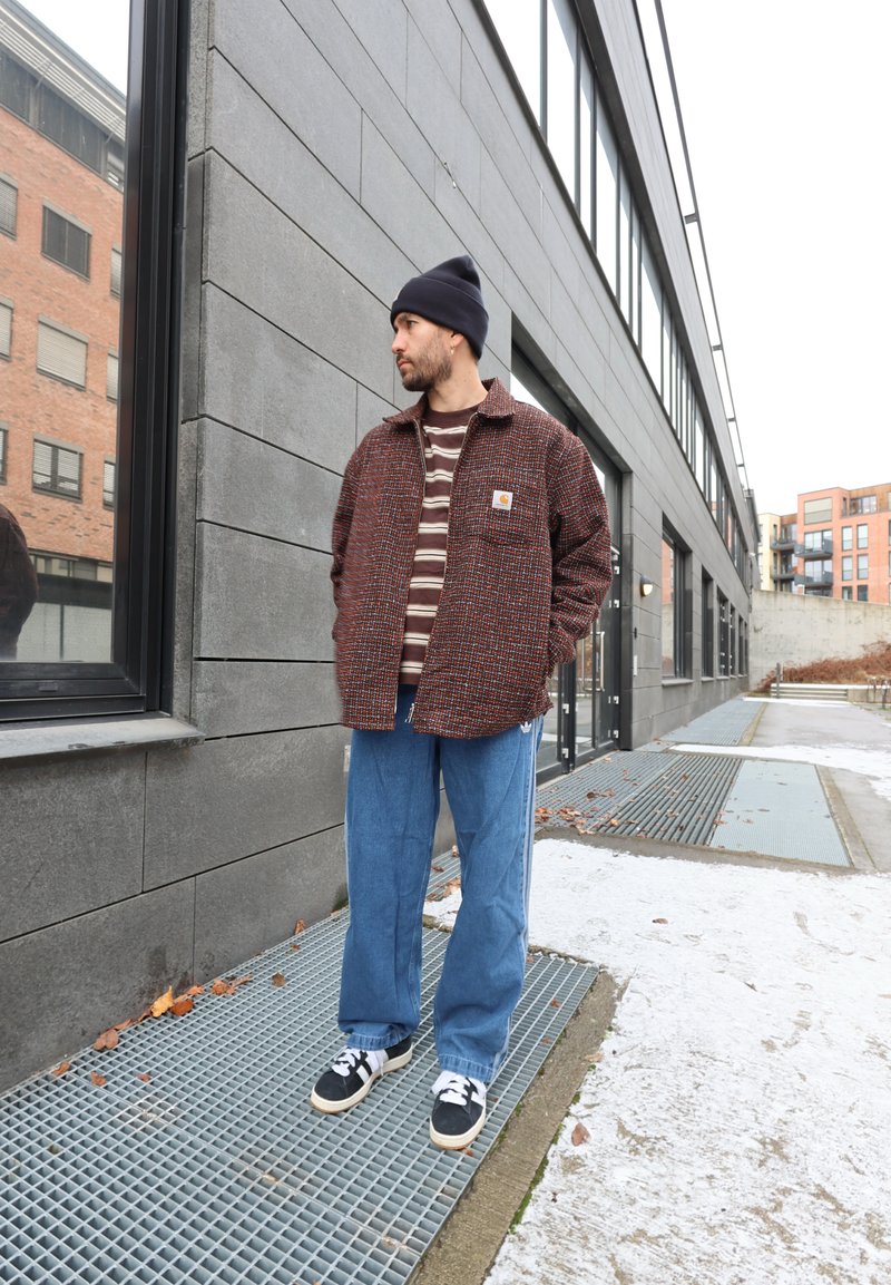 Veste texturée de couleur rouille avec une poche avant, portée sur une chemise à rayures marron et blanches. Jean bleu ample avec des accents blancs. Baskets noires.