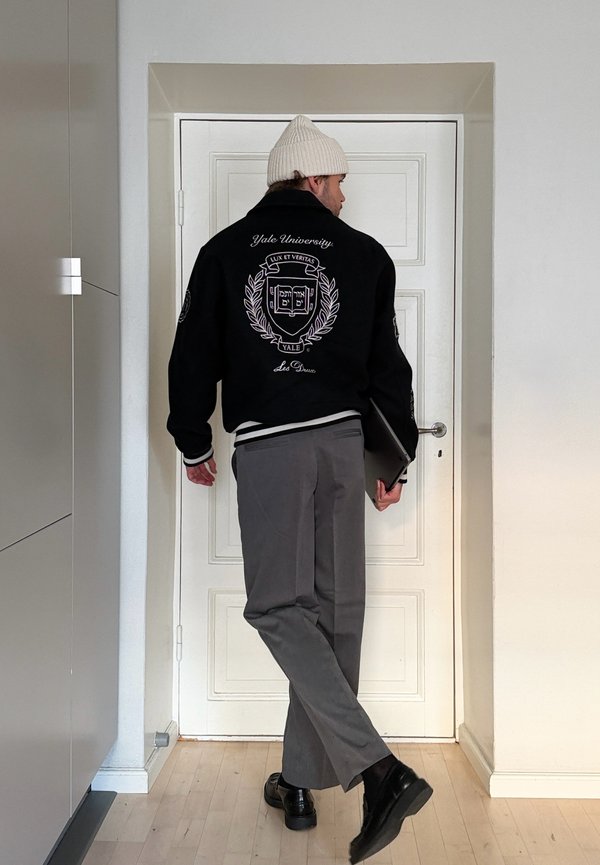 Homme portant une veste de l'université Yale et un bonnet beige, tenant un ordinateur portable, debout devant une porte blanche dans une pièce minimaliste.