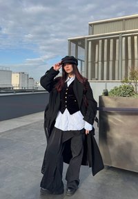 Jeune femme vêtue d'une tenue superposée noir et blanc avec manteau et casquette, debout sur une terrasse sur le toit près d'un grand bac à plantes sous un ciel nuageux.