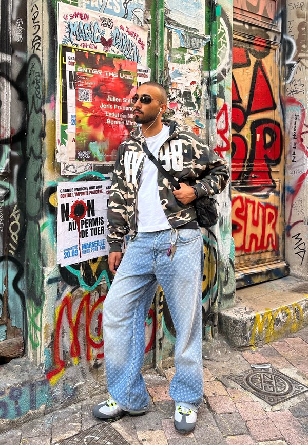 Hombre con chaqueta de camuflaje, vaqueros azul claro y gafas de sol, de pie frente a una pared cubierta de grafitis con carteles y arte urbano.