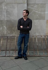 Schwarzes, langärmeliges Hemd mit Streifen, blaue Jeans und schwarze Schuhe. Steht vor einer grauen Wand mit einem rostigen Metallgeländer im Vordergrund.