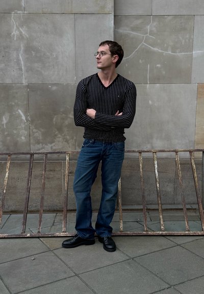 Camisa de manga larga con rayas negras, vaqueros azules y zapatos negros. De pie frente a una pared gris con una barandilla metálica oxidada en primer plano.